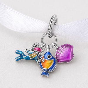Può includere: Un charm in argento con un subacqueo, un pesce e una conchiglia. Il subacqueo è blu con occhiali rosa. Il pesce è arancione e blu. La conchiglia è viola. Il charm è appeso a un nastro bianco.