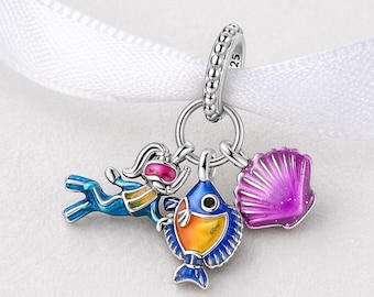 Puede incluir: Un charm de plata con un buzo, un pez y una concha. El buzo es azul con gafas rosas. El pez es naranja y azul. La concha es morada. El charm cuelga de una cinta blanca.