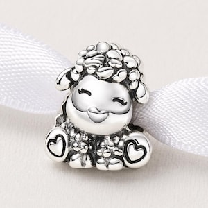 May include: A silver sheep charm with a smiling face and heart details. The sheep has a fluffy head and small ears. The charm is presented on a white ribbon.