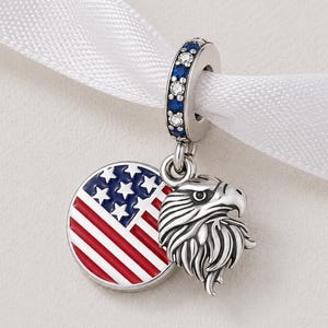 Op de afbeelding: Een zilveren bedel met een Amerikaans vlagontwerp en een gedetailleerde adelaarskop. De vlag is rood, wit en blauw, met een cirkel van sterren. De bedel hangt aan een ring versierd met blauwe en heldere stenen.
