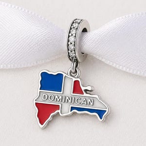 May include: A silver charm depicting the Dominican Republic's map. The charm is coloured with the national flag's colours: red, white, and blue. The word "DOMINICAN" is printed on the charm. It hangs from a silver loop with clear stones.