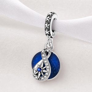 May include: A silver charm with a celestial theme. The charm features a dark blue circular pendant with a starburst design and a sparkling accent. The top loop has star cutouts. The charm is suspended from a white ribbon.