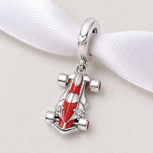 Peut inclure: Un charm en argent en forme de voiture de course, avec des détails en émail rouge et blanc. Le charm a quatre roues et deux pierres précieuses transparentes. Il est suspendu à une boucle en argent avec un cœur.