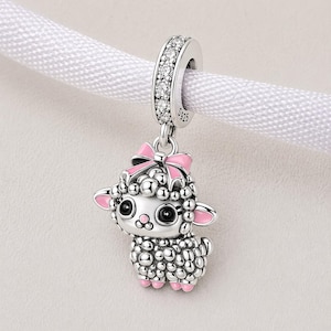 May include: A silver charm in the shape of a sheep, adorned with a pink bow and details. The charm has a rounded, textured body, pink ears and feet, and large black eyes. The top loop is set with clear stones.