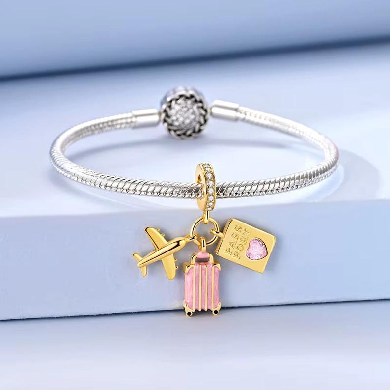 May include: A silver bracelet with a charm featuring a gold airplane, a pink suitcase, and a gold passport with a pink heart. The bracelet has a decorative clasp.