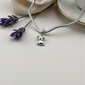Puede incluir: Un collar con colgante plateado con un colgante de vaca de las Tierras Altas. El dije, con detalles de cuernos y pelaje, cuelga de una cadena de serpiente. Ramas de lavanda moradas y un fondo blanco completan la composici&oacute;n.