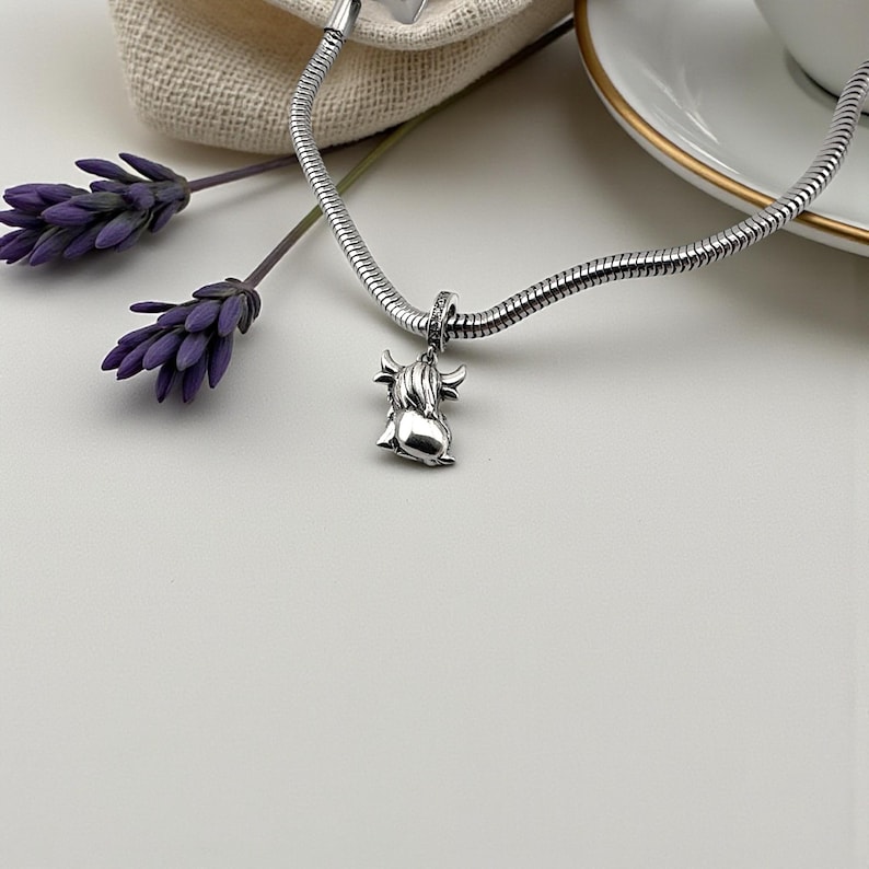 Puede incluir: Un collar con colgante plateado con un colgante de vaca de las Tierras Altas. El dije, con detalles de cuernos y pelaje, cuelga de una cadena de serpiente. Ramas de lavanda moradas y un fondo blanco completan la composici&oacute;n.
