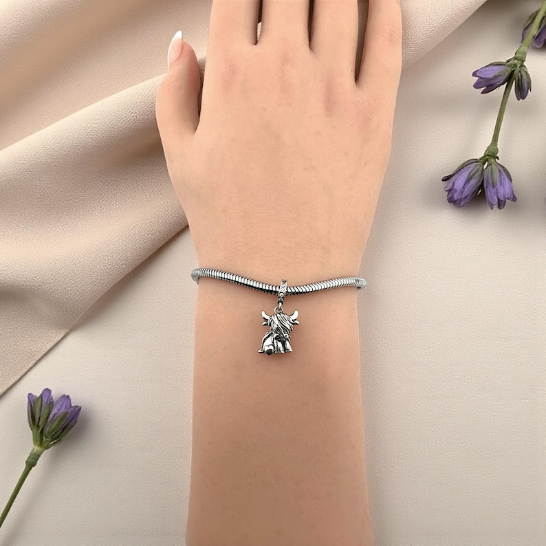 Puede incluir: Una pulsera con dije plateada con un dije de vaca de las Tierras Altas. El dije es detallado y cuelga de una pulsera de cadena de serpiente. La pulsera se usa en una mu&ntilde;eca, con un fondo suave y neutro.