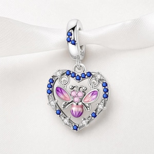 May include: A silver charm with a heart-shaped pendant. The heart is outlined with blue and clear crystals and features a bee with purple and pink wings. The charm has a loop with blue crystals for attaching to a bracelet or necklace.