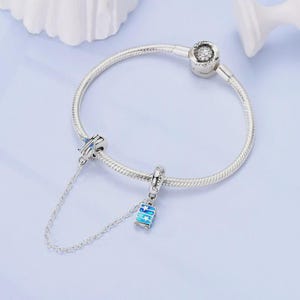 May include: A silver charm bracelet with two travel-themed charms. One charm is a blue airplane with blue accents, and the other is a blue suitcase charm with the word "Travel" on it. The bracelet is on a light blue surface.