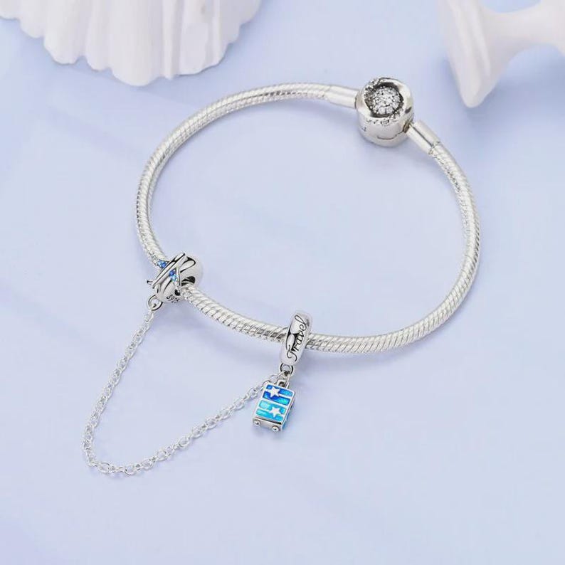May include: A silver charm bracelet with two travel-themed charms. One charm is a blue airplane with blue accents, and the other is a blue suitcase charm with the word "Travel" on it. The bracelet is on a light blue surface.