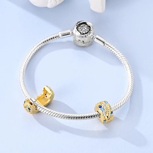 Puede incluir: Una pulsera de cadena de serpiente plateada con dos abalorios dorados y un cierre plateado. Un abalorio es dorado con piedras azules y transparentes, el otro es dorado con piedras azules. El cierre es plateado con un dise&ntilde;o floral negro.