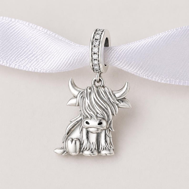 Puede incluir: Un charm de plata con un dise&ntilde;o de vaca de las Tierras Altas. El charm presenta una representaci&oacute;n detallada de la vaca con cuernos y pelo largo. El lazo superior est&aacute; incrustado con peque&ntilde;as piedras transparentes y cuelga de una cinta blanca.