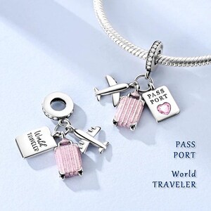 May include: Silver travel-themed charm with a suitcase, airplane, and passport design. The pink suitcase charm has a textured surface. The passport charm has a heart-shaped gem. The text "World Traveler" and "Pass Port" are visible.