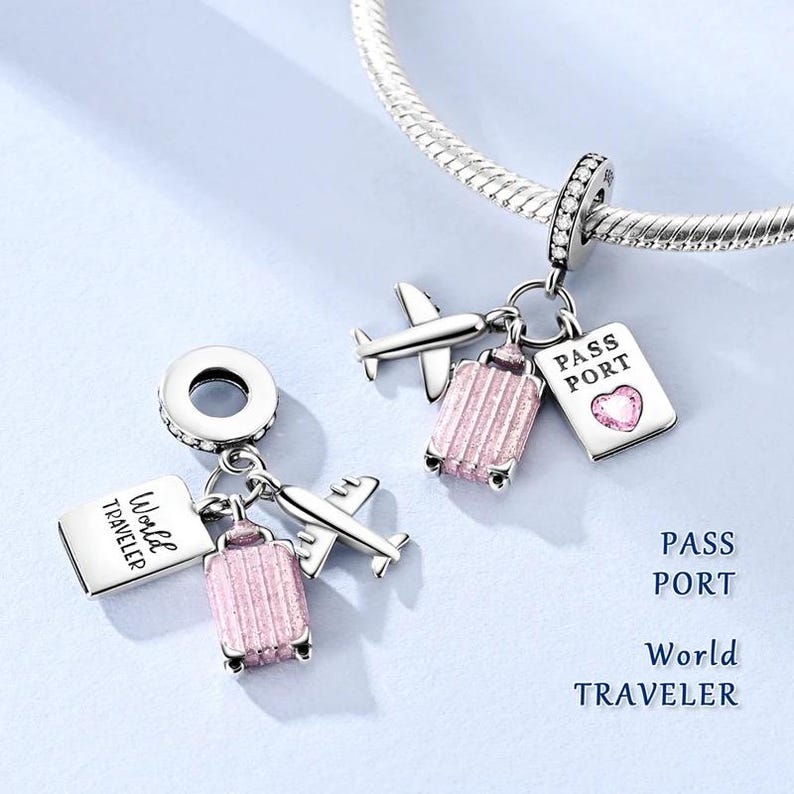 May include: Silver travel-themed charm with a suitcase, airplane, and passport design. The pink suitcase charm has a textured surface. The passport charm has a heart-shaped gem. The text "World Traveler" and "Pass Port" are visible.