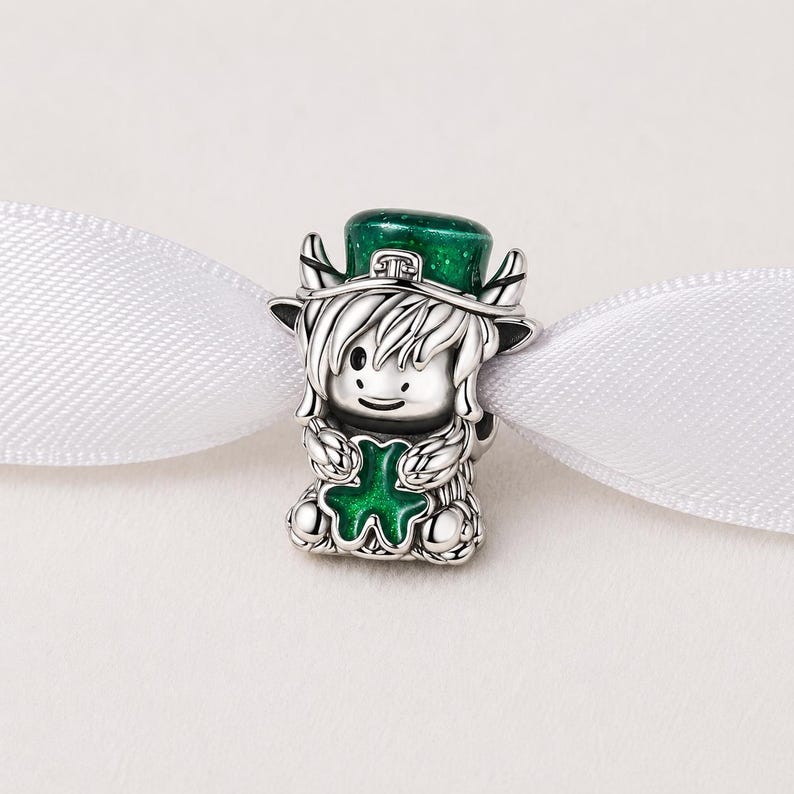 May include: A silver charm featuring a cartoon leprechaun with a green hat and a green four-leaf clover. The leprechaun has silver hair and is holding the clover. The charm is on a white ribbon.