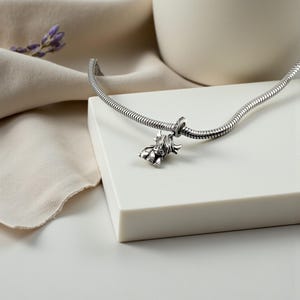Puede incluir: Una pulsera de plata con un charm de vaca de las Tierras Altas. El charm es detallado y tiene una apariencia texturizada. La pulsera est&aacute; sobre una superficie blanca con un fondo neutro. Una ramita de lavanda est&aacute; en el fondo.