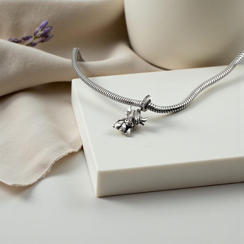 Puede incluir: Una pulsera de plata con un charm de vaca de las Tierras Altas. El charm es detallado y tiene una apariencia texturizada. La pulsera est&aacute; sobre una superficie blanca con un fondo neutro. Una ramita de lavanda est&aacute; en el fondo.