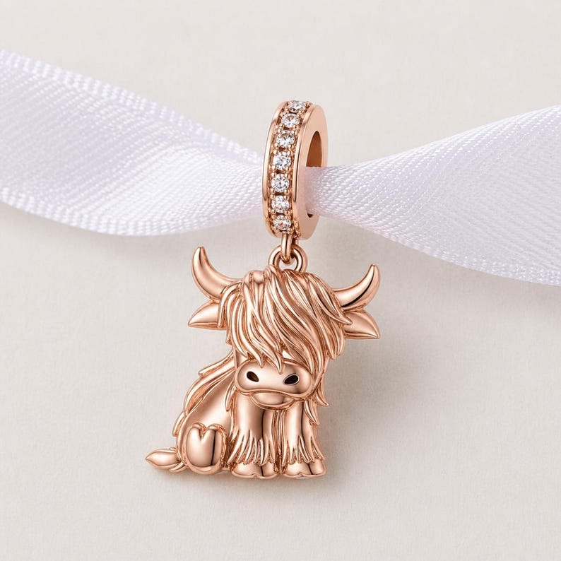 Puede incluir: Un charm de oro rosa con un dise&ntilde;o de vaca de las Tierras Altas. El charm tiene una apariencia detallada y texturizada con una figura de vaca sentada. El lazo superior est&aacute; incrustado con peque&ntilde;as piedras preciosas transparentes. El charm est&aacute; suspendido de una cinta blanca.