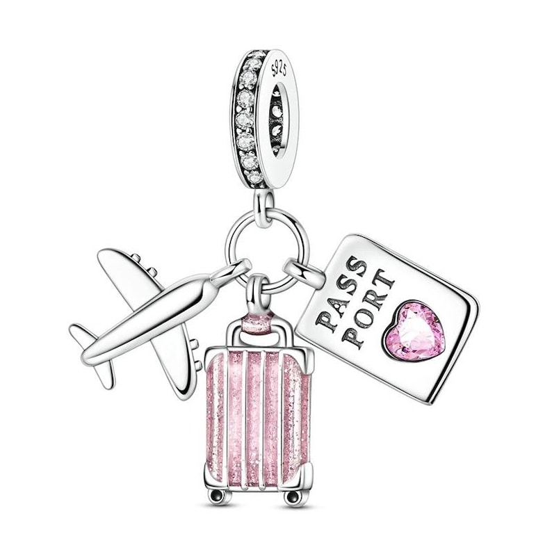 May include: A silver charm with travel-themed elements. It features a small airplane, a pink glitter suitcase, and a passport with a heart-shaped gem. The charm is suspended from a ring with clear, sparkling accents.