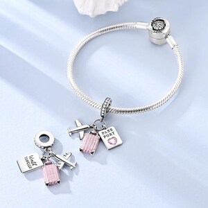 May include: A silver charm bracelet with travel-themed charms. Charms include a pink suitcase, airplane, and passport with the text "PASS PORT" and a heart. The bracelet is on a light blue surface.