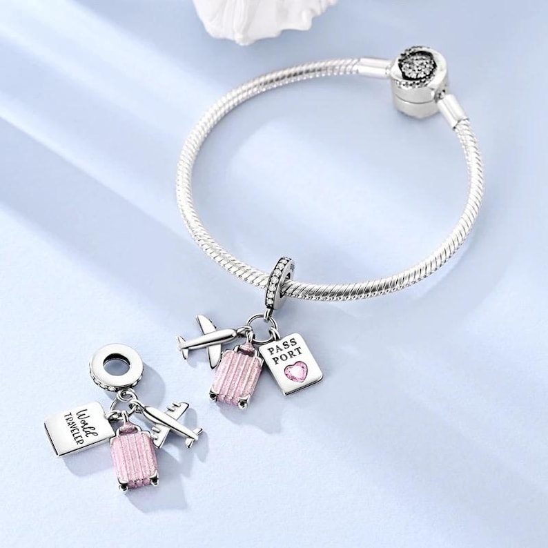 May include: A silver charm bracelet with travel-themed charms. Charms include a pink suitcase, airplane, and passport with the text "PASS PORT" and a heart. The bracelet is on a light blue surface.