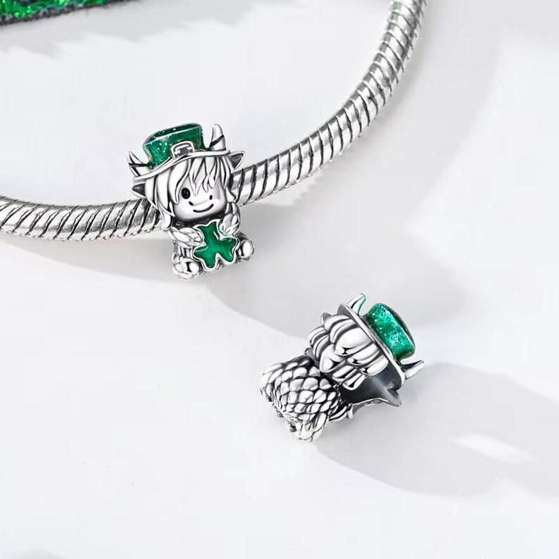 May include: Two silver charms featuring a cartoon Highland cow wearing a green hat and holding a green shamrock. The charms are detailed with black and white accents and are designed to be used as a bracelet charm.