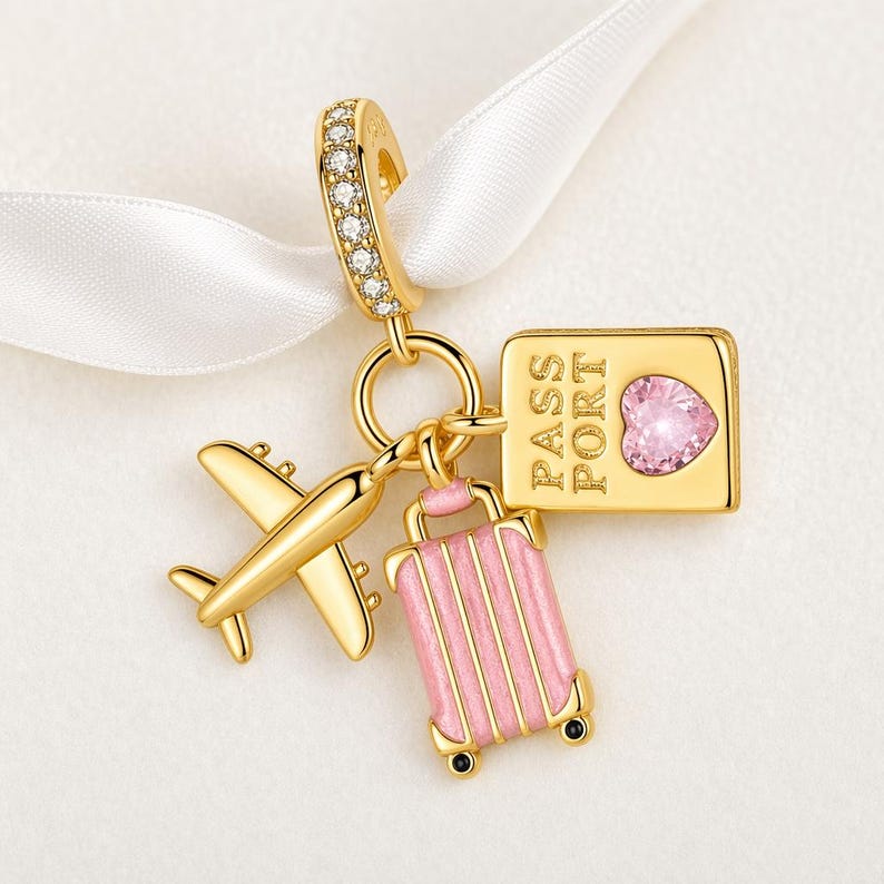 May include: A gold-colored charm with travel-themed elements. It features a small airplane, a pink suitcase, and a passport with a heart-shaped gem. The charm is suspended from a gold loop with clear stones.