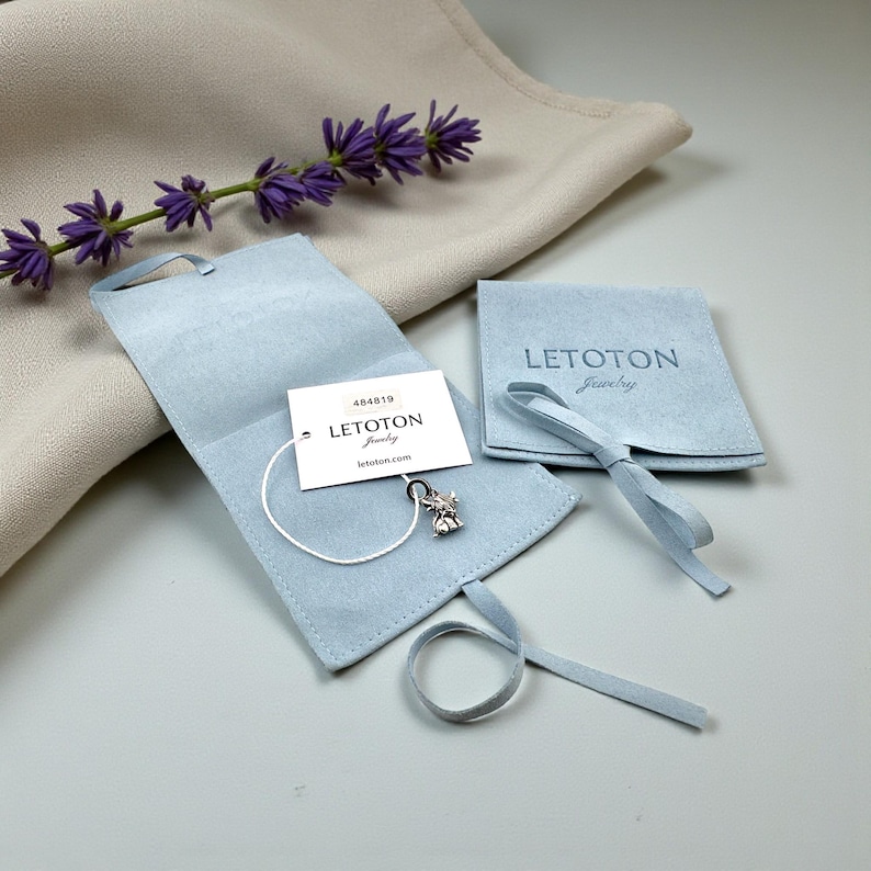 Puede incluir: Una bolsa de joyer&iacute;a azul claro con una cinta a juego, un peque&ntilde;o dije de plata y una etiqueta que dice "LETOTON Jewelry". Una ramita de lavanda p&uacute;rpura descansa sobre una tela beige.