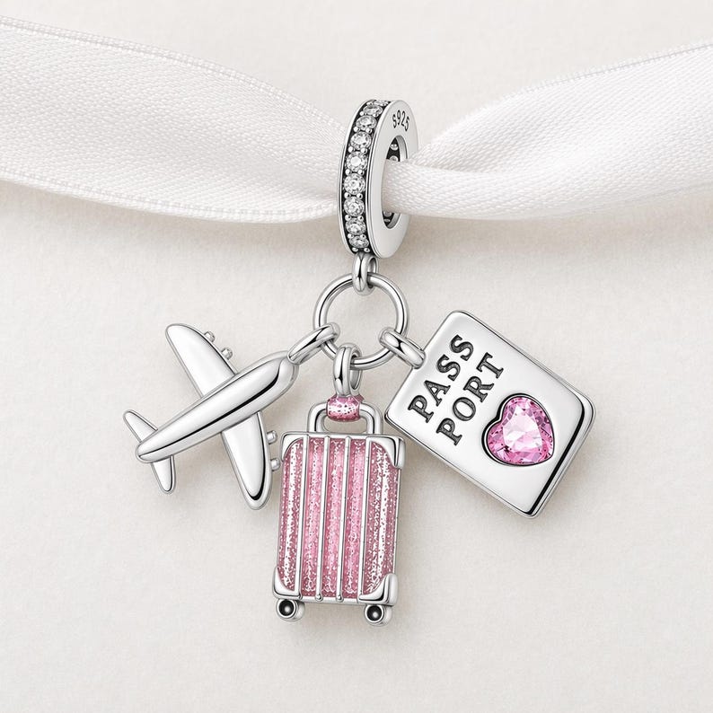 May include: A silver charm with travel-themed elements. It features a small airplane, a pink glitter suitcase, and a passport with a heart-shaped gem. The charm is suspended from a ring with clear stones.