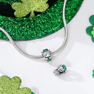 May include: Two silver charms with a St. Patrick's Day theme. One charm features a smiling leprechaun with a green hat and shamrock. The other charm is a close-up of the leprechaun's hat. A silver chain and green shamrock decorations are also visible.