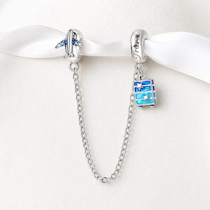 May include: A silver charm bracelet with two travel-themed charms. One charm is an airplane with blue accents, and the other is a blue suitcase with star details. The charms are connected by a silver chain.