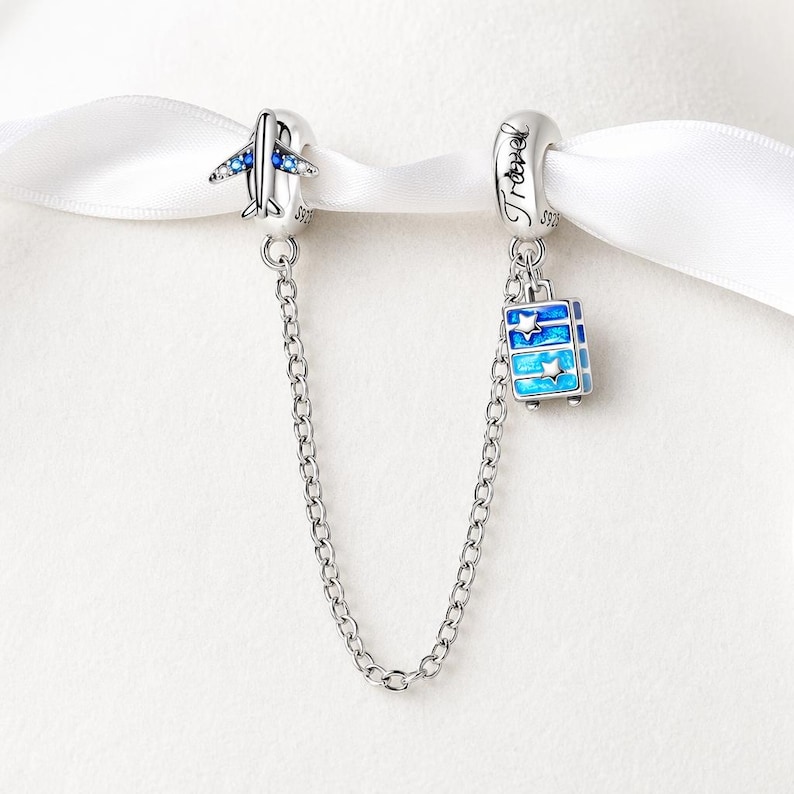 May include: A silver charm bracelet with two travel-themed charms. One charm is an airplane with blue accents, and the other is a blue suitcase with star details. The charms are connected by a silver chain.