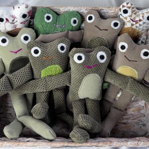 May include: A collection of handmade frog plush toys in a woven basket. The frogs have large white eyes, stitched smiles, and various shades of green and brown. Some have patterned details, and all have long arms and legs.
