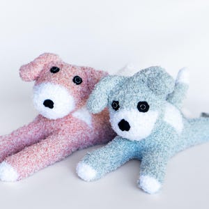 May include: Two plush dog toys in a lying down position. One is pink with white accents, the other is blue with white accents. Both have black eyes and noses. The toys are made of a soft, textured material.