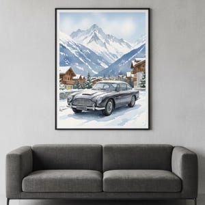 May include: Watercolor print of a classic gray car parked in a snowy landscape with snow-capped mountains and chalets. The artwork is framed in a black frame and hangs above a gray sofa.
