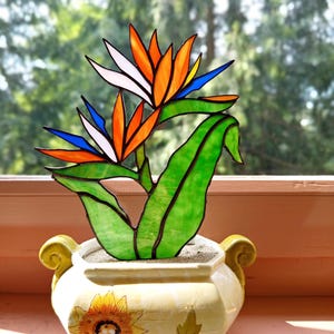 Può includere: Un fiore uccello del paradiso in vetro colorato con petali arancioni, bianchi e blu e foglie verdi, inserito in un vaso di ceramica color crema con un disegno di girasole dipinto sul davanti e due manici curvi sui lati.