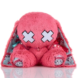May include: A pink plush bunny with white cross-shaped eyes and a beaded necklace. The inner ears and paws feature a floral and cat print. The bunny is a soft, cuddly toy.