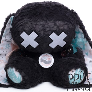 May include: Black plush bunny with white X's for eyes, a teal ribbon, and a circular tag with a hand design. The bunny is sitting on a white background with a pink and teal patterned fabric.