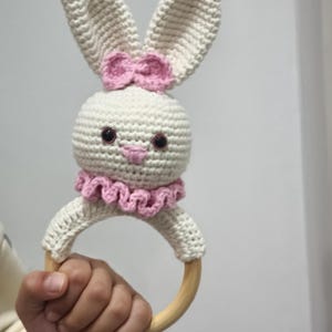 May include: A white crocheted bunny rattle with pink ears and a pink bow. The rattle has a wooden ring for a handle.