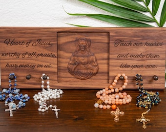 May include: A wooden plaque with carved religious imagery and text, holding several rosaries. The plaque features an image of Jesus and the phrases "Heart of Jesus" and "Touch our hearts." Rosaries in blue, white, peach, and black are displayed.