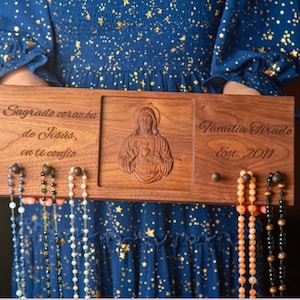 May include: A wooden rosary holder with engraved text and a carved image of Jesus. The holder displays several rosaries with various bead colors. The text includes "Sagrado corazon de Jesus, en ti confio" and "Familia Tirado Est. 2011".