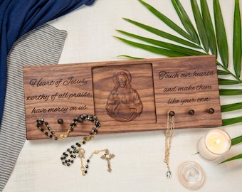 May include: A wooden wall-mounted rosary holder with engraved text and a carved image of Jesus. The holder has three pegs for hanging items and is accompanied by a rosary and a necklace. A lit candle and a palm frond are also visible.