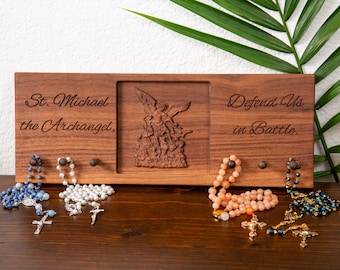 May include: A wooden wall hanging with the text "St. Michael the Archangel, Defend Us in Battle." It features a carved image of St. Michael and holds several rosaries with crosses. The rosaries are in shades of blue, white, and orange.