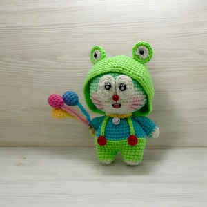 May include: A green crocheted stuffed toy with a frog hat, blue overalls, and red buttons. The toy is holding a colorful balloon string.