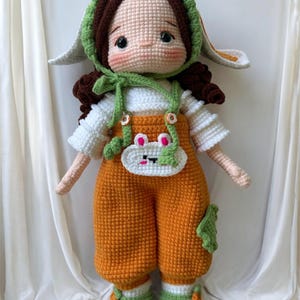 May include: A handmade crochet doll with brown hair, wearing an orange jumpsuit, white shirt, and green hat with bunny ears. The doll has rosy cheeks and a small white bib with a bunny face. The doll is approximately 18 inches tall.