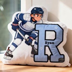 May include: A plush, white pillow in the shape of a hockey player, wearing a blue and white uniform. The pillow has a large, light blue letter "R" with the name "Ryan" and a hockey puck. The player is holding a hockey stick.