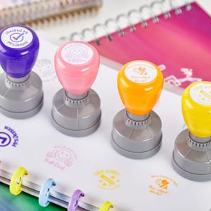 Pode incluir: Conjunto de cinco carimbos de professor coloridos com mensagens diferentes. Os carimbos são feitos de plástico e têm uma base cinzenta. As mensagens nos carimbos incluem "Checked by Mrs. Jessica", "Bee-utiful Work!", "Good Work!", "It's All Good!" e "Keep it Up!"