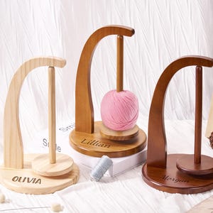 May include: Three wooden yarn holders in varying shades of brown. The holders feature a curved top and a central dowel for holding yarn. The base of each holder is engraved with a name: Olivia, Lillian, and Morgan. A pink ball of yarn is on the middle holder.