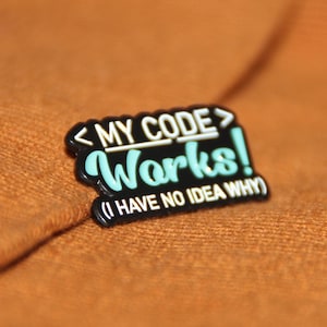 以下が含まれることがあります： 白文字で「<MY CODE> Works! (I HAVE NO IDEA WHY)」と書かれた黒いエナメルピン。 「Works!」の文字は薄い青色です。
